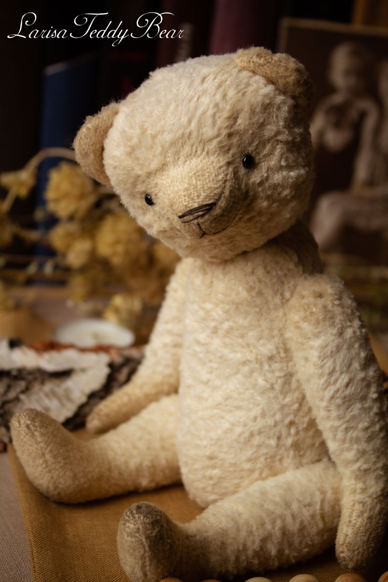 May include: A cream-colored teddy bear with a stitched nose and black eyes sits on a brown surface. The bear has a fluffy, worn texture. The text "Larisa Teddy Bear" is visible in the top left corner of the image.