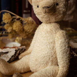 May include: A cream-colored teddy bear with a stitched nose and black eyes sits on a brown surface with wooden beads. The bear has a fluffy, soft texture. The text "Larisa Teddy Bear" is visible in the background.