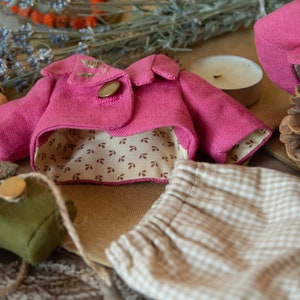 Pu&ograve; includere: Una giacca di tessuto rosa con fodera a fantasia marrone e bianca, un paio di pantaloni a quadri beige e bianchi e una piccola borsa di tessuto verde.