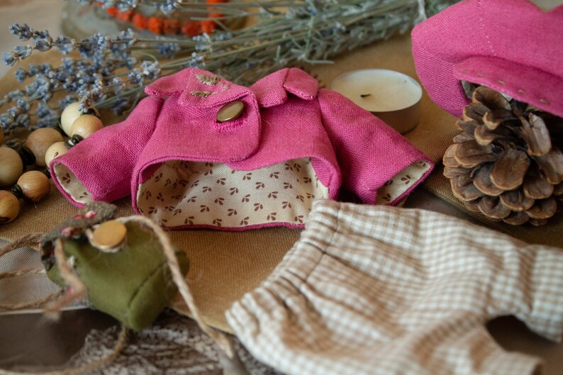 Pu&ograve; includere: Una giacca di tessuto rosa con fodera a fantasia marrone e bianca, un paio di pantaloni a quadri beige e bianchi e una piccola borsa di tessuto verde.