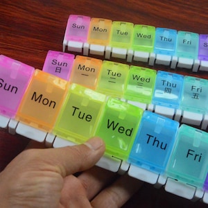 Può includere: Un portapillole settimanale con sette scomparti, ognuno etichettato con un giorno della settimana. Gli scomparti sono di vari colori, tra cui rosa, arancione, giallo, verde, blu e viola. I giorni sono in inglese.
