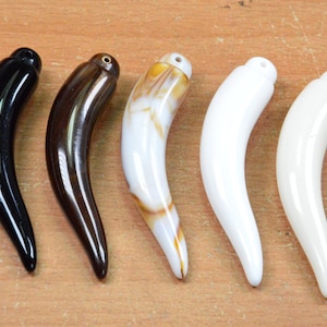 May include: Five different colored tooth-shaped pendants. The pendants are made of a smooth, shiny material and have a hole at the top for hanging. The colors are black, brown, white with brown swirls, white, and off-white.