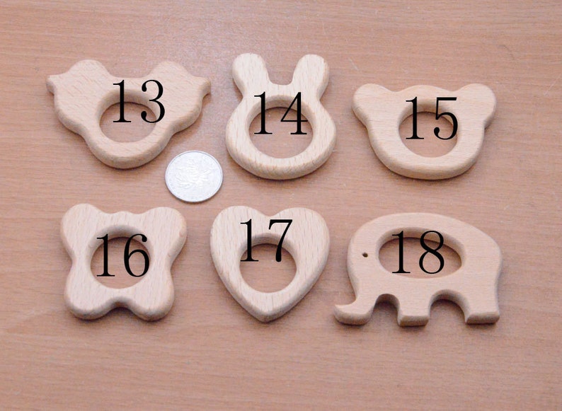 May include: A collection of six wooden baby teethers in various shapes: a butterfly, bunny, bear, flower, heart, and elephant. Each teether has a smooth, natural wood finish and a central hole for easy gripping. A coin is included for size reference.