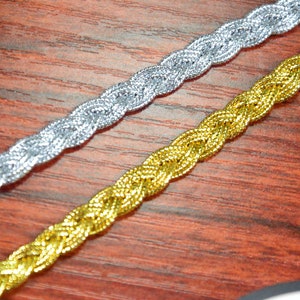 May include: Two braided trims, one silver and one gold, are shown on a wooden surface. The trims are both woven with a similar pattern.
