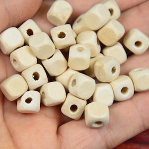 Puede incluir: Primer plano de una mano sosteniendo un montón de cuentas cuadradas de madera natural con agujeros en el centro. Las cuentas están sin terminar y listas para ser pintadas o teñidas.