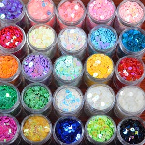 May include: A collection of small, clear jars filled with colorful sequins. The sequins are in various colors, including pink, blue, green, yellow, and purple. The jars are arranged on a wooden surface.