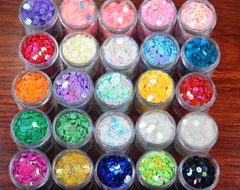 May include: A collection of small, clear jars filled with colorful sequins. The sequins are in various colors, including pink, blue, green, yellow, and purple. The jars are arranged on a wooden surface.