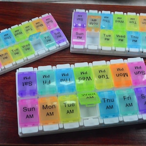 May include: Three colorful pill organizers with clear, labeled compartments for each day of the week, AM and PM. The lids are in various colors, including purple, blue, green, yellow, and orange. The organizers sit on a wooden surface.