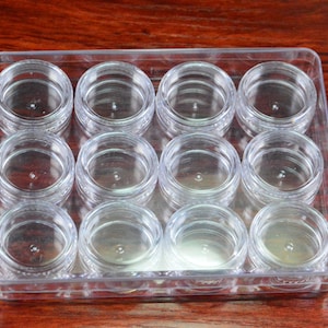 Può includere: Una scatola di contenimento in plastica trasparente con dodici piccoli contenitori rotondi trasparenti. I contenitori sono disposti in una griglia 3x4.