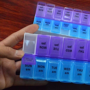 May include: Two clear plastic pill organizers, one held by a hand. Each organizer has two rows of compartments labeled with days of the week and "am" and "pm". The compartments are blue and purple.