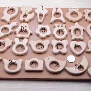 May include: A collection of twenty-four wooden baby teethers in various shapes, including animals, flowers, and geometric designs. The teethers are made of light-colored wood and are arranged on a wooden surface. A coin is included for size reference.