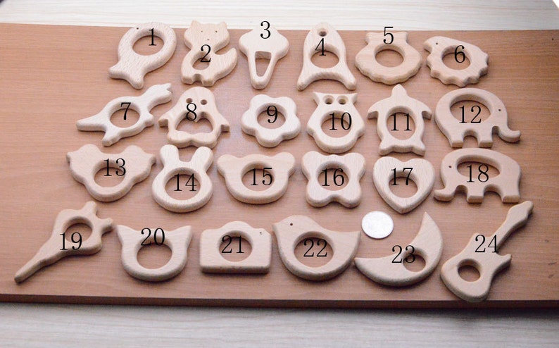 May include: A collection of twenty-four wooden baby teethers in various shapes, including animals, flowers, and geometric designs. The teethers are made of light-colored wood and are arranged on a wooden surface. A coin is included for size reference.