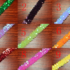 May include: Nine different colors of sequined trim, each color is shown in a separate row. The trim is made of small, round sequins that are sewn together in a continuous line. The sequins are iridescent and reflect light, creating a shimmering effect. The trim is available in a variety of colors, including green, purple, blue, pink, yellow, orange, red, and white.