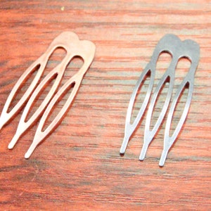 May include: Two metal hair combs. One is copper-colored, the other is silver. Each comb has three tines and a decorative top. The combs are displayed on a dark wood surface.