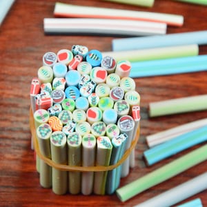 May include: A collection of colorful, patterned polymer clay sticks for nail art. The sticks feature various Christmas-themed designs, including Santa Claus, Christmas trees, and candy canes. The sticks are arranged in a bundle and secured with a rubber band.