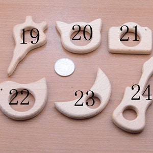 May include: A collection of six unfinished wooden baby teethers in various shapes: a flower, a cat, a camera, a bird, a crescent moon, and a guitar. Each teether has a hole in the center. The teethers are made of light-colored wood.