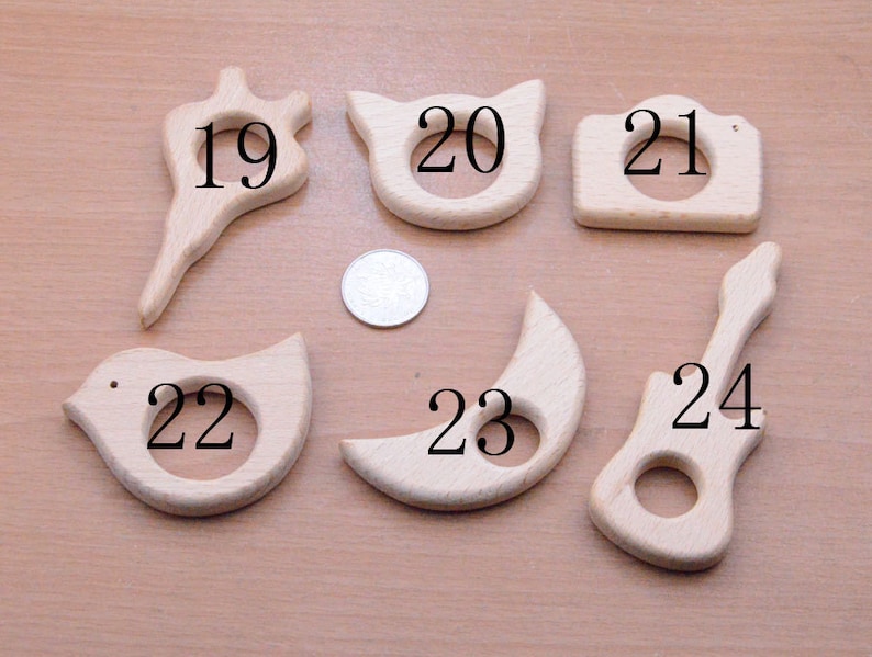 May include: A collection of six unfinished wooden baby teethers in various shapes: a flower, a cat, a camera, a bird, a crescent moon, and a guitar. Each teether has a hole in the center. The teethers are made of light-colored wood.