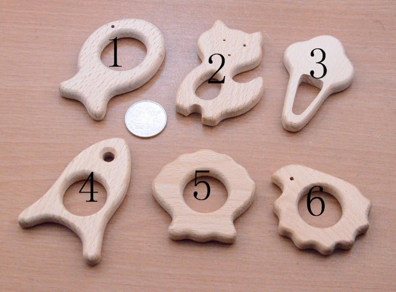 May include: A collection of six unfinished wooden baby teethers in various shapes, including a fish, cat, and seashell. Each teether has a hole for easy gripping. The teethers are made of light-colored wood.