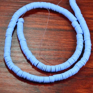 May include: A strand of light blue, flat disc beads, strung together on a thin, clear thread. The beads are arranged in a circular pattern, with a portion of the strand overlapping. The background is a dark wood surface.
