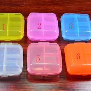 May include: Six small, square pill boxes with lids. The boxes are numbered 1 through 6 and are in different colours: yellow, pink, blue, clear, light pink, and orange.
