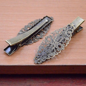 May include: Two antique bronze-colored hair clips. Each clip features an ornate, openwork design with floral and scroll details. The clips have a classic barrette style with a hinged mechanism for securing hair.