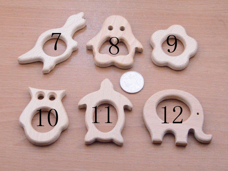 May include: A collection of six wooden baby teethers in various animal shapes, including a bird, penguin, flower, owl, turtle, and elephant. Each teether has a circular opening in the center and is made of light-colored wood.