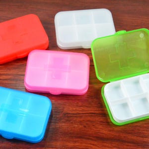Puede incluir: Una colección de pastilleros coloridos en rojo, blanco, rosa, azul y verde. Cada estuche rectangular tiene un símbolo de cruz y una tapa con bisagras. El estuche verde abierto revela seis compartimentos.