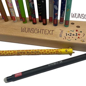 May include: A wooden pen holder with various pens and styluses. The holder has the words "WUNSCHTEXT" and "WUNSCHBILD" engraved on it, along with a math equation and a drawing. One pen has a giraffe pattern, and another says "BE CURIOUS".