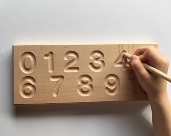May include: Wooden tracing board with numbers 0-10 carved into the surface. A child's hand is tracing the number 4 with a wooden stylus.