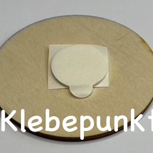 May include: A round wooden disc with a white adhesive circle in the center. The text "Klebepunkt" is printed on the bottom of the disc.