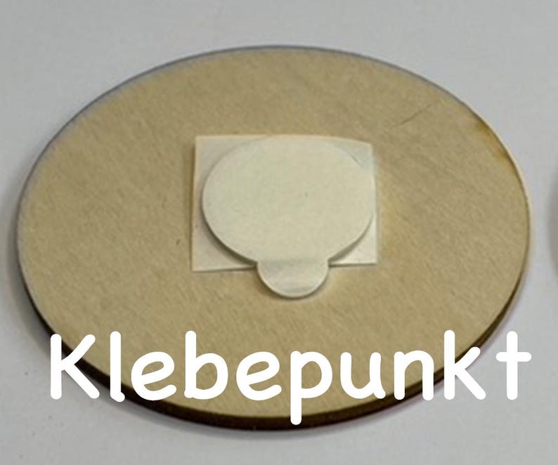 May include: A round wooden disc with a white adhesive circle in the center. The text "Klebepunkt" is printed on the bottom of the disc.