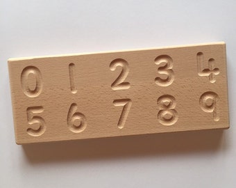 May include: Wooden number tracing board with the numbers 0 through 9 carved into the surface. The board is light brown and has a smooth finish.