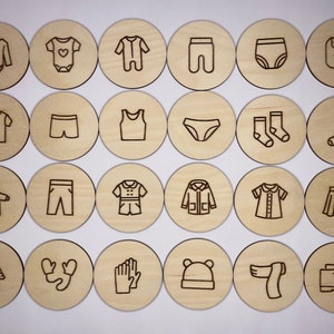 May include: Set of 20 wooden circles with different baby clothing items engraved on them. The items include a onesie, a shirt, pants, a skirt, a bib, socks, mittens, a hat, and a scarf.