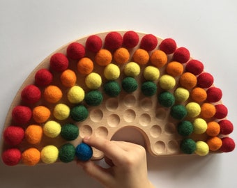 May include: A wooden toy shaped like a half-circle with 36 holes. The holes are filled with colourful felt balls in a rainbow pattern. A hand is holding a blue felt ball.