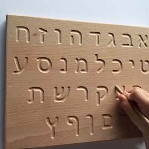 May include: A wooden board with Hebrew alphabet letters carved into it. A child's hand is tracing the letters with a wooden stylus.