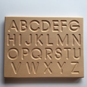 May include: Wooden alphabet tracing board with uppercase letters A through Z carved into the surface. The board is light brown and has a smooth finish.