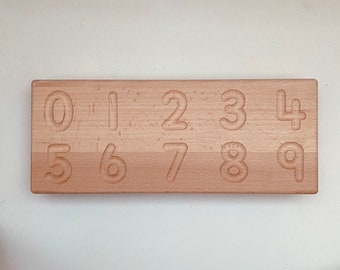 May include: A wooden number tracing board with the numbers 0 through 9 carved into the surface. The board is rectangular and made of light-colored wood. The numbers are in a sans-serif font and are indented into the wood.