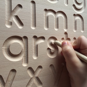May include: A wooden alphabet tracing board with raised letters. A child's hand is tracing the letter 's' with a wooden stylus.