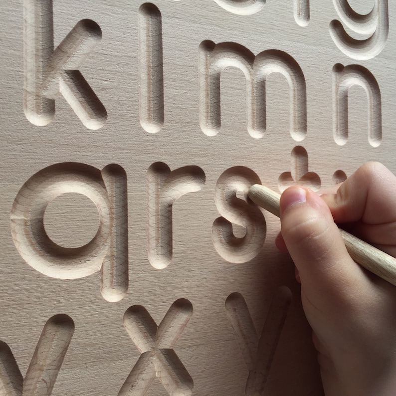 May include: A wooden alphabet tracing board with raised letters. A child's hand is tracing the letter 's' with a wooden stylus.