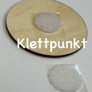May include: A round wooden disc with a white adhesive velcro dot attached to the top. The disc has the word "Klettpunkt" written on it. A separate white adhesive velcro dot is shown on a clear plastic backing.