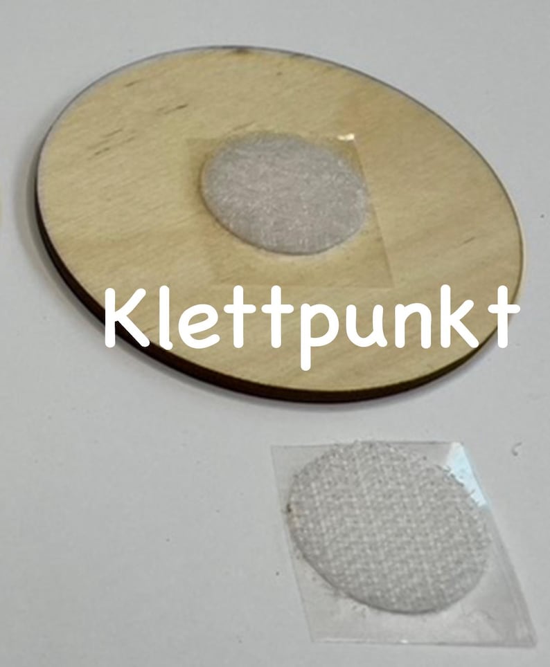 May include: A round wooden disc with a white adhesive velcro dot attached to the top. The disc has the word "Klettpunkt" written on it. A separate white adhesive velcro dot is shown on a clear plastic backing.