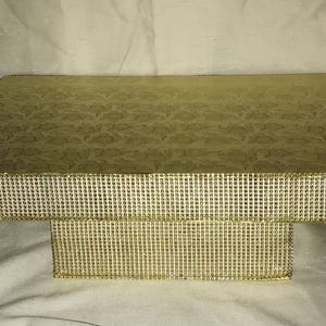 May include: A two-tiered gold cake stand with a decorative, embossed top and a rhinestone trim. The stand is perfect for displaying cakes, cupcakes, or other desserts.