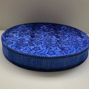 May include: A round, blue fabric-covered box with a floral pattern on the top. The sides are a darker blue, textured fabric. The box is sitting on a white surface.