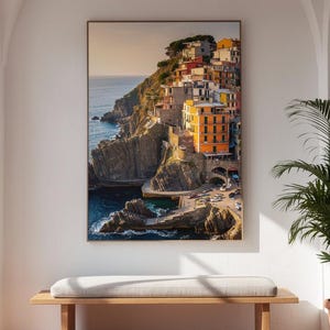 Può includere: Una stampa incorniciata di un villaggio costiero italiano con edifici colorati su una scogliera. L'opera d'arte è appesa sopra una panca di legno con un cuscino chiaro. Una pianta in vaso è a destra e la stanza ha porte ad arco e pavimento piastrellato.