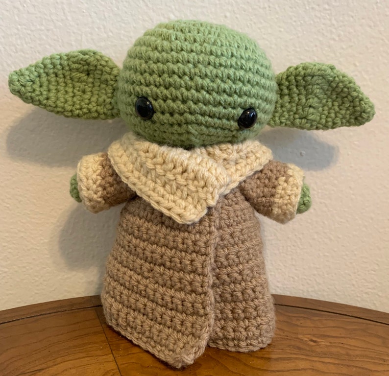 May include: A handmade crochet doll of a character with large ears, green skin, and black eyes. The doll is wearing a tan and brown robe. The doll is approximately 8 inches tall.