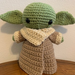 May include: A handmade crochet doll of a character with large ears and black eyes. The doll is primarily green, with a tan robe and a cream-colored collar. The doll is standing on a wooden surface.