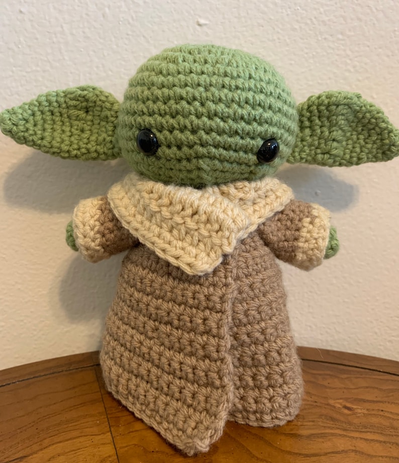 May include: A handmade crochet doll of a character with large ears and black eyes. The doll is primarily green, with a tan robe and a cream-colored collar. The doll is standing on a wooden surface.