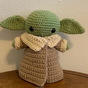 May include: A handmade crochet doll of a character with large ears, wearing a tan coat with a light tan collar. The doll's head and ears are green, and it has black eyes. The doll is approximately 8 inches tall.