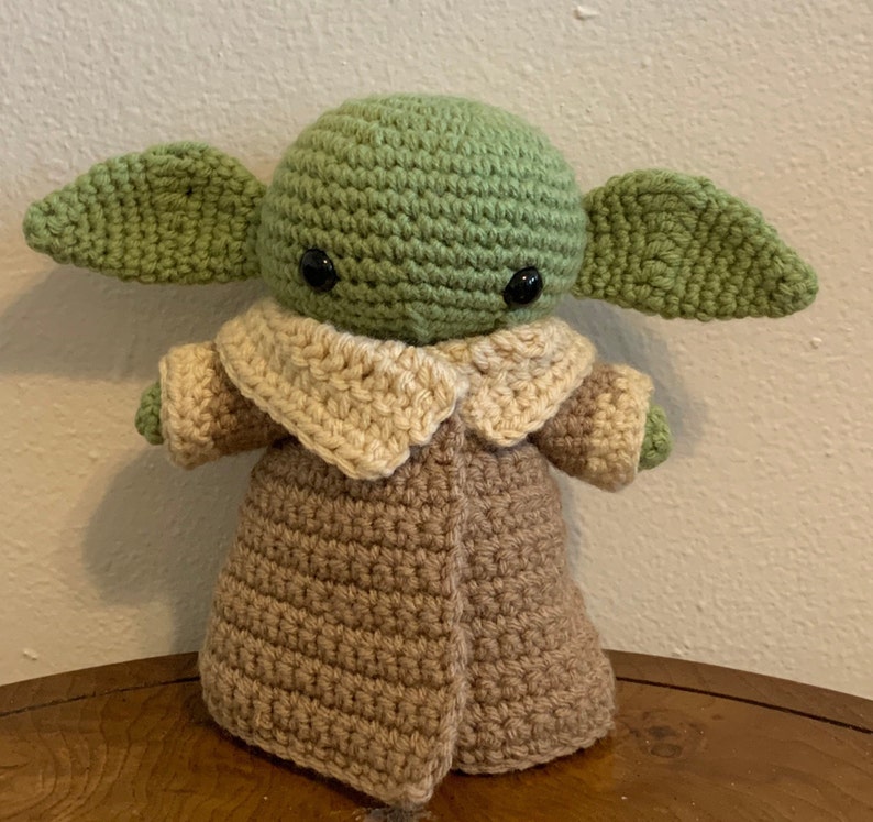 May include: A handmade crochet doll of a character with large ears, wearing a tan coat with a light tan collar. The doll's head and ears are green, and it has black eyes. The doll is approximately 8 inches tall.