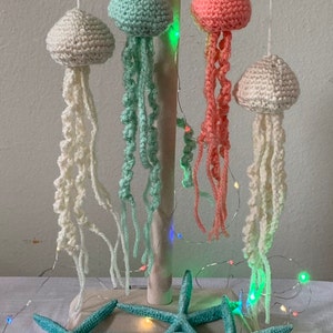 May include: Handmade crochet jellyfish ornaments in cream, seafoam green, and coral pink, suspended from a wooden stand. The jellyfish have long, flowing tentacles. Teal starfish are arranged at the base, with string lights adding a festive touch. A whimsical, nautical theme.